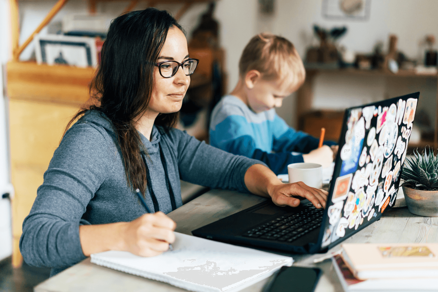 Stressed female entrepreneur sitting at desk with laptop and notebook, overwhelmed by marketing tasks